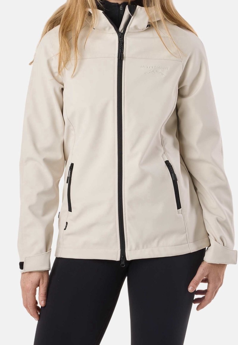Femme portant une veste zippée beige clair avec deux poches zippées et un pantalon noir, vue des épaules aux hanches.