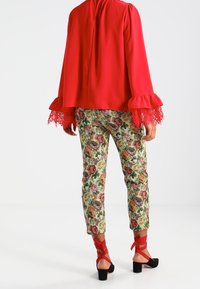 Blusa rossa a maniche lunghe con dettagli in pizzo, abbinata a pantaloni con motivo floreale caratterizzati da rose verdi e multicolori e sandali neri con tacco a blocco.