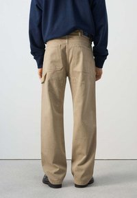 Persona vestida con pantalones de trabajo beige salpicados de pintura y zapatos negros, de espaldas, con una camisa de manga larga azul marino sobre un fondo liso.
