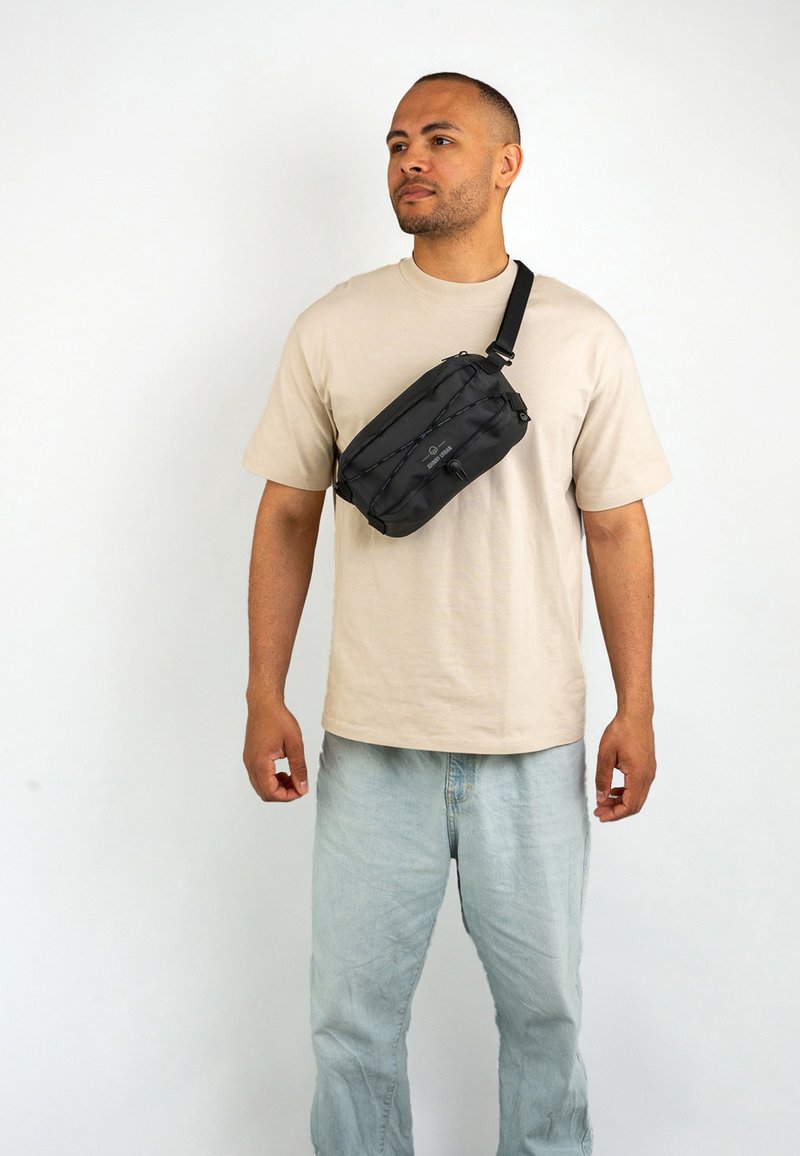 Czarna torba na ramię z teksturowaną powierzchnią, zamek błyskawiczny, regulowany pasek i detal z elastycznego sznurka, noszona na beżowej koszuli.