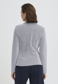 Veste grise cintrée à manches longues, col tailleur, tissu texturé et détails de coutures, associée à un pantalon bleu marine. Vue de dos.