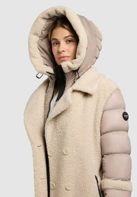 Bēša mākslīgās kažokādas coat ar apjomīgu, pūkīgu kapuci; ar lieliem pogām un quilted detaļām uz piedurknēm. Ar rāvējslēdzēju un regulējamiem elementiem.