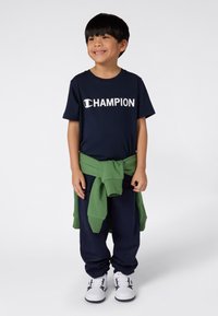 Tamna mornarsko plava majica s bijelim logotipom "CHAMPION", u paru s tamnim tajicama i zelenom duksericom zavezanom oko struka, nosi se s bijelim cipelama.