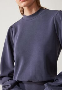 Haut à manches longues bleu marine avec un col montant, présentant une texture lisse et des manches froncées, offrant un design ajusté et minimaliste.