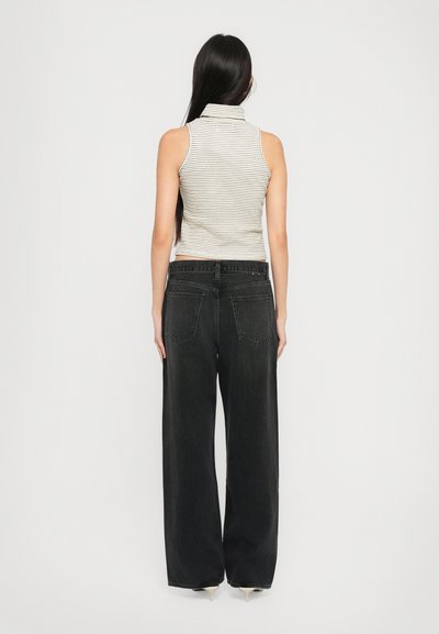 Eine Frau mit langen, dunklen Haaren, die ein ärmelloses gestreiftes Rollkragenoberteil, hochgeschnittene, breite schwarze Jeans und weiße High Heels trägt, steht mit dem Rücken zur Kamera.