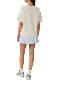T-shirt en coton crème surdimensionné à manches courtes, associé à une jupe plissée bleu clair. Le modèle porte des baskets argentées avec des chaussettes blanches.