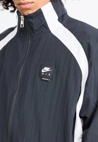 Jaqueta Nike AIR, preta com riscas brancas laterais, gola alta, fecho com zíper, tecido texturizado, apresenta um patch com a marca no peito.