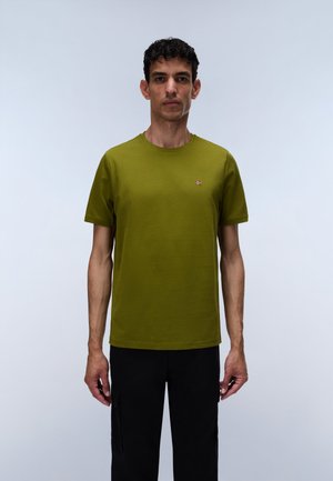 Homme portant un t-shirt à manches courtes vert olive avec un petit logo sur la poitrine, debout devant un fond gris clair uni.
