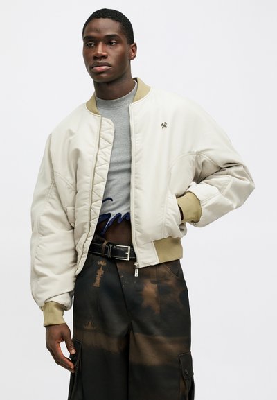 Jachetă bomber crem cu guler și manșete ribbate. Prezintă un design matlasat, închidere cu fermoar pe față și un emblemă brodată pe piept.