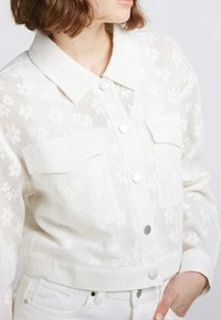 Veste blanche courte en tissu de dentelle avec broderies florales, dotée d'un col pointu, de deux poches poitrine et de boutons en métal argenté.
