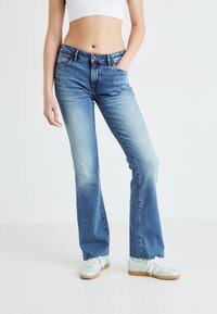 Jeans acampanados de color azul claro con textura desgastada, cinco bolsillos y cierre con botón en la parte superior. Combinados con zapatillas ligeras para un look casual.