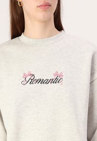 Felpa grigia con testo ricamato in nero e rosa "Romantic" e fiocchi decorativi. Materiale confortevole, scollatura a costine e design classico.