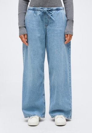 Osoba nosící světlé modré široké džíny s elastickým pasem na stahovací šňůrku, šedý žebrovaný svetr a bílé tenisky.