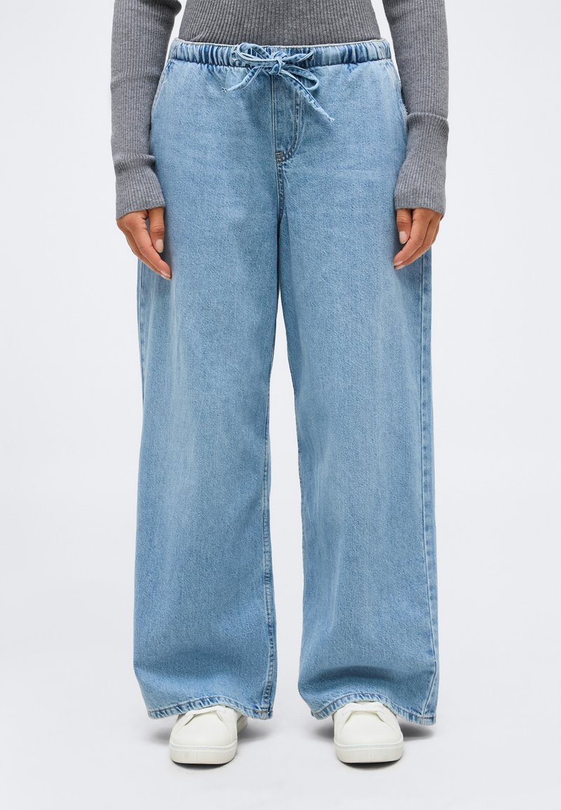 Άτομο που φοράει ανοιχτό μπλε τζιν με φαρδιά άκρα και ελαστική μέση με κορδόνι, γκρι ριγέ πουλόβερ και λευκά αθλητικά παπούτσια.