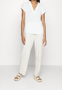 Vit kortärmad topp med v-ringning, i kombination med ljusbeiga vida byxor och krämfärgade sandaler, mot en enfärgad bakgrund.