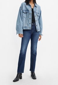 Lichtblauwe spijkerjas met twee zakken aan de voorkant, zwarte top eronder, donkerblauwe spijkerbroek en zwarte enkellaarzen, staand tegen een witte achtergrond.