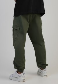 Olivgröna cargobyxor i bomull med elastiska muddar och sidofickor. Bärs med vita sneakers med grå detaljer och en enkel design.