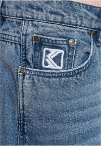 Farkut vaaleansinisellä pesulla, joissa on valkoinen K-logo-patch takataskussa, ja korostettu kultaisilla metallinapeilla.