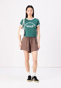 Green cropped t-shirt with white trim and graphic text, paired with brown drawstring shorts. Black sneakers with white and beige accents.
