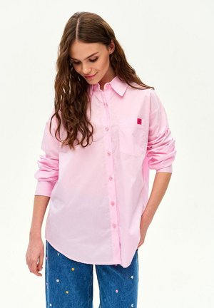 Kobieta z długimi brązowymi włosami, ubrana w luźną jasnoróżową koszulę na guziki i niebieskie jeansy z drobnymi, kolorowymi haftowanymi kwiatami.