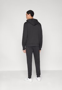 Σετ μαύρο φούτερ με κουκούλα και παντελόνι από λείο ύφασμα. Το φούτερ διαθέτει κορδόνι ρύθμισης και τσέπη καγκουρό. Το σύνολο ολοκληρώνεται με λευκά αθλητικά παπούτσια.