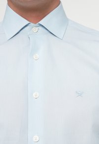 Lyseblå skjorte med klassisk krage, med hvite knapper og en subtil brodert logo på venstre side. Glatt tekstur.