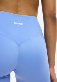 Κοντινό πλάνο ενός ατόμου που φοράει ανοιχτό μπλε ψηλόμεσα κολάν με το κείμενο «STRONGER» στη μέση, πάνω σε λευκό φόντο.