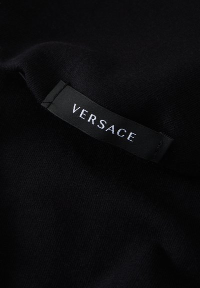 Fekete szövet sima textúrával; feltűnő fekete címkével, amelyen fehér nagybetűkkel olvasható: "VERSACE".