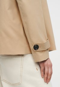 Μανσέτα μπεζ σακακιού με μαύρη λεπτομέρεια κουμπιού που φοριέται πάνω από κρεμ παντελόνι, εμφανίζοντας την πίσω τσέπη και το χέρι που ακουμπάει στο πλάι.