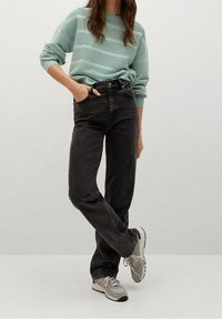 Personne portant un pull rayé vert menthe, un jean noir taille haute et des baskets grises, debout avec une jambe croisée sur l'autre.