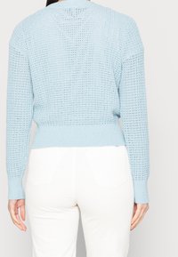 Vaaleansininen neulepusero, jossa on avoin kuvio, pitkät hihat, resorikärjet ja -helma, yhdistettynä valkoisiin teksturoituihin housuihin.