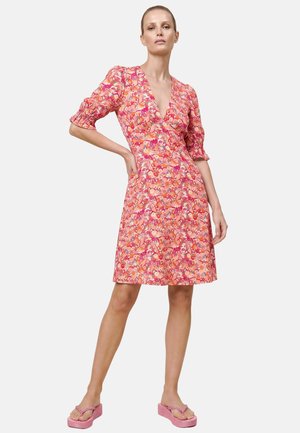 Femme portant une robe fleurie rose et orange longueur genou avec des manches trois-quarts bouffantes et des tongs compensées roses, debout, une main sur la hanche.