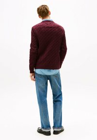 Burgundy cable-knit sweater with long sleeves, ribbed cuffs, and hem, paired with light blue denim jeans and black footwear, viewed from the back.
