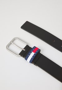 Schwarzer Leder-Gürtel mit einer silbernen rechteckigen Schnalle. Hat ein gestreiftes Stoffakzent in Blau, Rot und Weiß in der Nähe der Schnalle.