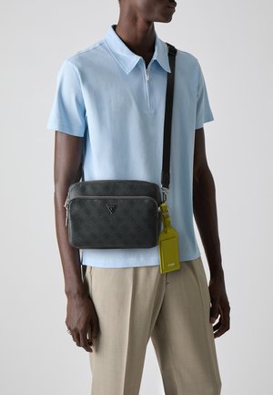 Sac bandoulière noir à motifs avec ferrures argentées, fermetures éclair et une étiquette jaune vif. Le mannequin porte un polo bleu clair et un pantalon beige.