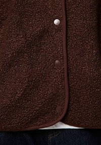 Primer plano de chaqueta de forro polar marrón texturizado con botones a presión y ribete marrón oscuro sobre vaqueros azules.