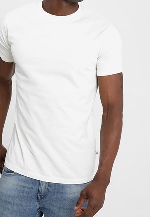 Mann trägt ein schlichtes weißes T-Shirt mit kurzen Ärmeln und Rundhalsausschnitt sowie blaue Jeans, steht mit einer Hand nahe der Hosentasche vor einem hellen Hintergrund.