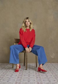 Mujer con cabello rubio sentada en una silla de madera, vistiendo un suéter rojo con mangas recortadas, jeans azules anchos y tacones rojos sobre un suelo estampado.