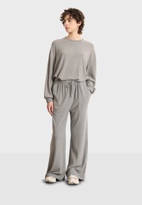 Persona in piedi che indossa pantaloni della tuta grigi larghi, una felpa abbinata oversize e sneakers bianche su uno sfondo chiaro e semplice.