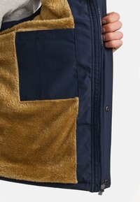 Chaqueta azul marino con forro de terciopelo marrón y un bolsillo lateral. Presenta un cierre de cremallera y botones a presión. Textura suave en el interior.