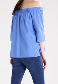 Top blu off-the-shoulder con maniche a tre quarti, realizzato in tessuto leggero, con scollo arricciato e fessure laterali. Abbinato a jeans.