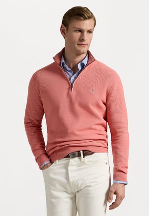 Pullover corallo lavorato a maglia con colletto a zip. Indossato sopra una camicia a righe blu e bianche. Abbinato a pantaloni bianchi e una cintura marrone.