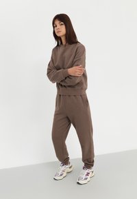 Sweatshirt en coton marron et joggers assortis avec des poignets côtelés. Baskets blanches avec des accents violets et une semelle épaisse. Fond neutre.