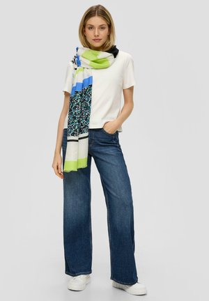 Écharpe multicolore avec un mélange de motifs, principalement en vert, bleu et noir. Portée par-dessus une chemise blanche, avec des bords frangés. Jean en denim.