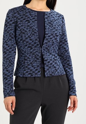 Nainen pukeutuneena pitkähihainen, istuva laivastonsininen ja musta kuvioitu bleiseri mustan paidan päällä, yhdistettynä mustiin housuihin neutraalilla taustalla.