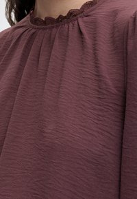 Femme portant un chemisier violet texturé avec un col froncé et une bordure en dentelle visible au-dessus du col.
