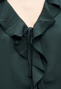 Πράσινο σκούρο μπλούζα με V-λαιμόκοψη και υφασμάτινη υφή, που διαθέτει στρωματοποιημένες βολάν και λεπτό δεσίμα στον λαιμό.