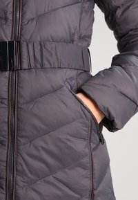 Padded gray coat with a quilted design, featuring a glossy black belt, zip pockets, and a high collar. Textured fabric and contrasting seams.