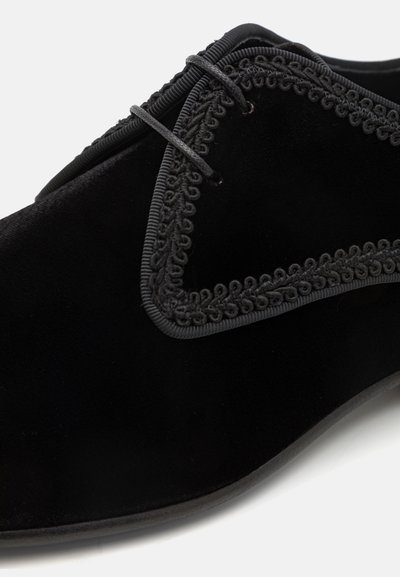 Chaussure en velours noir avec un bord cranté texturé et un design à lacets. Surface lisse avec des lacets noirs et des détails de couture subtils.