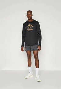 Tricou negru cu mânecă lungă, având un logo auriu în formă de munte, asortat cu pantaloni scurți gri cu un model. Modelul poartă adidași și șosete albe.